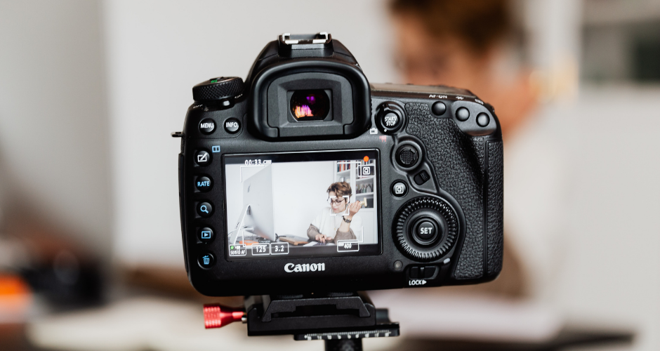 Professional DSLR camera recording a person speaking at a desk, capturing a corporate interview-style video for marketing purposes.