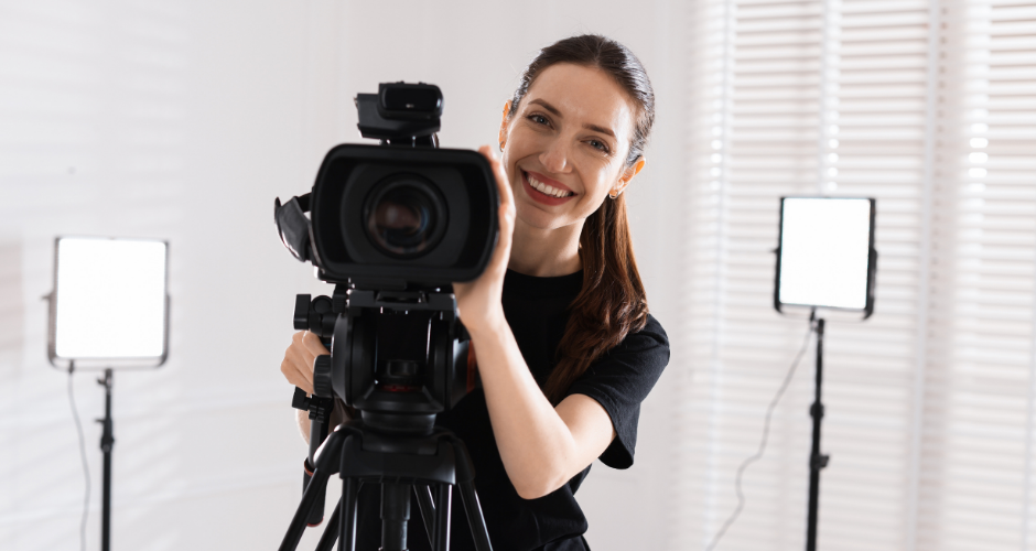 Smiling female videographer adjusting a professional video camera in a bright studio with soft lighting equipment.