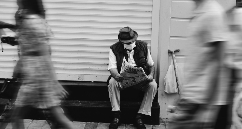 Black-and-white photo of a seated man reading a newspaper while blurred pedestrians pass by.